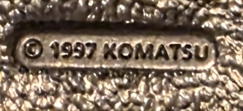 Op de afbeelding: Close-up van een metalen object met de tekst "&copy; 1997 KOMATSU" in reli&euml;f. Het object heeft een getextureerd oppervlak en lijkt een kleine, rechthoekige plaat of embleem te zijn.