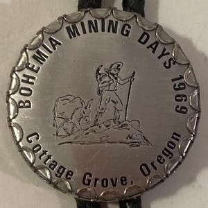 Pode incluir: Gravata bolo de prata com um pingente circular que apresenta o texto "Bohemia Mining Days 1969 Cottage Grove, Oregon". O pingente também representa um homem com um burro em uma paisagem montanhosa.