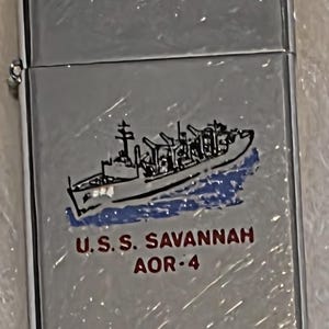 Puede incluir: Encendedor plateado con una ilustración detallada de un barco. La imagen incluye el texto "U.S.S. SAVANNAH AOR-4" en letras rojas, con un fondo azul y blanco.