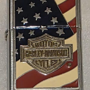 May include: A silver-colored lighter with a patriotic design. The top features the American flag with blue, white, and red colors. The Harley-Davidson logo is prominently displayed in the center.