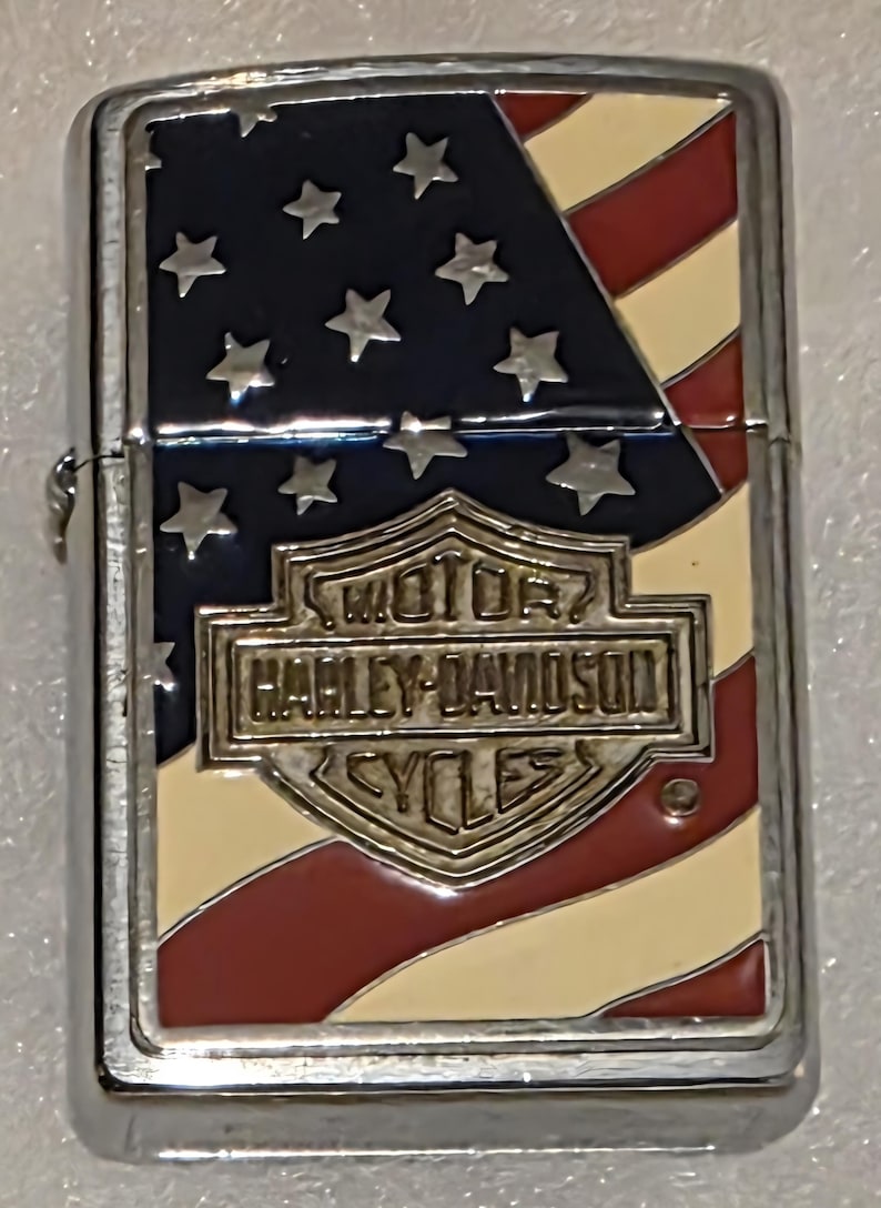May include: A silver-colored lighter with a patriotic design. The top features the American flag with blue, white, and red colors. The Harley-Davidson logo is prominently displayed in the center.