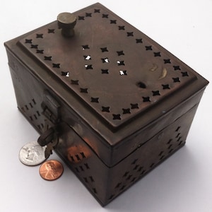 Puede incluir: Una pequeña caja rectangular de latón con una tapa con bisagras. La tapa tiene un patrón decorativo de estrellas recortadas y un pequeño pomo en la parte superior. La caja tiene una cerradura y una llave en el lateral.