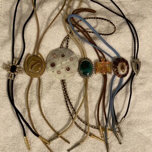 May include: A collection of bolo ties with various decorative pendants. The pendants include a cross, an oval with a shell design, a white circular pendant with pink accents, a green oval stone, a gold-colored bird, a brown and white oval, and a silver arrowhead.