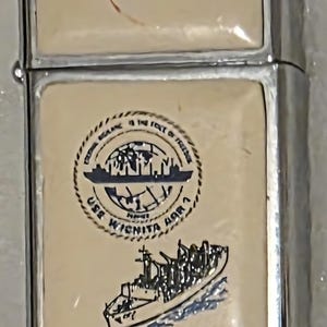 Può includere: Un accendino argentato con un pannello frontale color crema. Il pannello presenta una grafica blu navy e bianca di una nave e un emblema circolare con testo. Il testo "U.S.S. WICHITA AOR-1" è stampato in rosso.