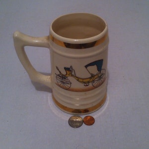 May include: A white ceramic beer stein with a gold rim and a yellow carriage with a blue top printed on the front.