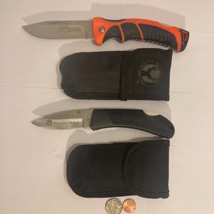 May include: Two folding knives with sheaths and a few coins. One knife has an orange and black handle, the other has a black handle. Both knives have silver blades and the text "Whitetails Unlimited" on the blade.