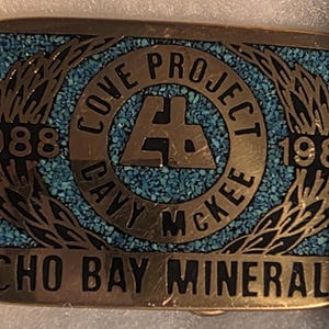 May include: Gold-coloured rectangular belt buckle with turquoise inlay. The buckle has the text "COVE PROJECT DAVY MCKEE" around a central logo, with the years "1988" and "1989". The bottom reads "ECHO BAY MINERALS".