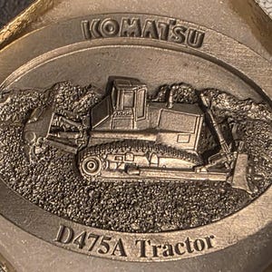 Op de afbeelding: Zilverkleurig Komatsu D475A Tractor embleem. Het embleem toont een gedetailleerd reli&euml;f van een bulldozer tegen een getextureerde achtergrond. Het woord "KOMATSU" staat bovenaan, "D475A Tractor" onderaan.