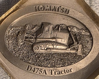 Puede incluir: Emblema plateado Komatsu D475A Tractor. Presenta un relieve detallado de una excavadora sobre un fondo texturizado. La palabra "KOMATSU" está en la parte superior y "D475A Tractor" en la inferior.
