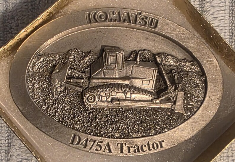 Op de afbeelding: Zilverkleurig Komatsu D475A Tractor embleem. Het embleem toont een gedetailleerd reli&euml;f van een bulldozer tegen een getextureerde achtergrond. Het woord "KOMATSU" staat bovenaan, "D475A Tractor" onderaan.