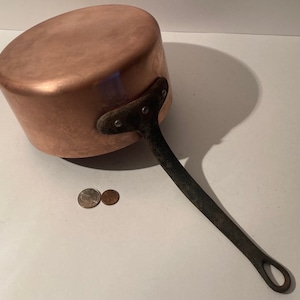 May include: A copper saucepan with a black iron handle. The saucepan is about 6 inches in diameter and has a rounded bottom. The handle is attached to the side of the saucepan with rivets.