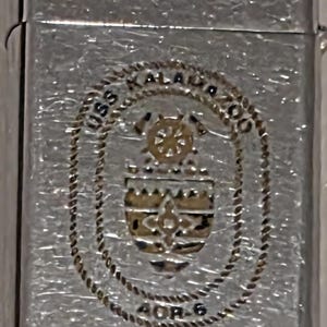 May include: Silver-toned metal lighter with a textured surface. The lighter features a detailed emblem with the text "USS KALAMAZOO" and "AOR-6" in a circular design. The emblem includes a ship's wheel and other nautical elements.