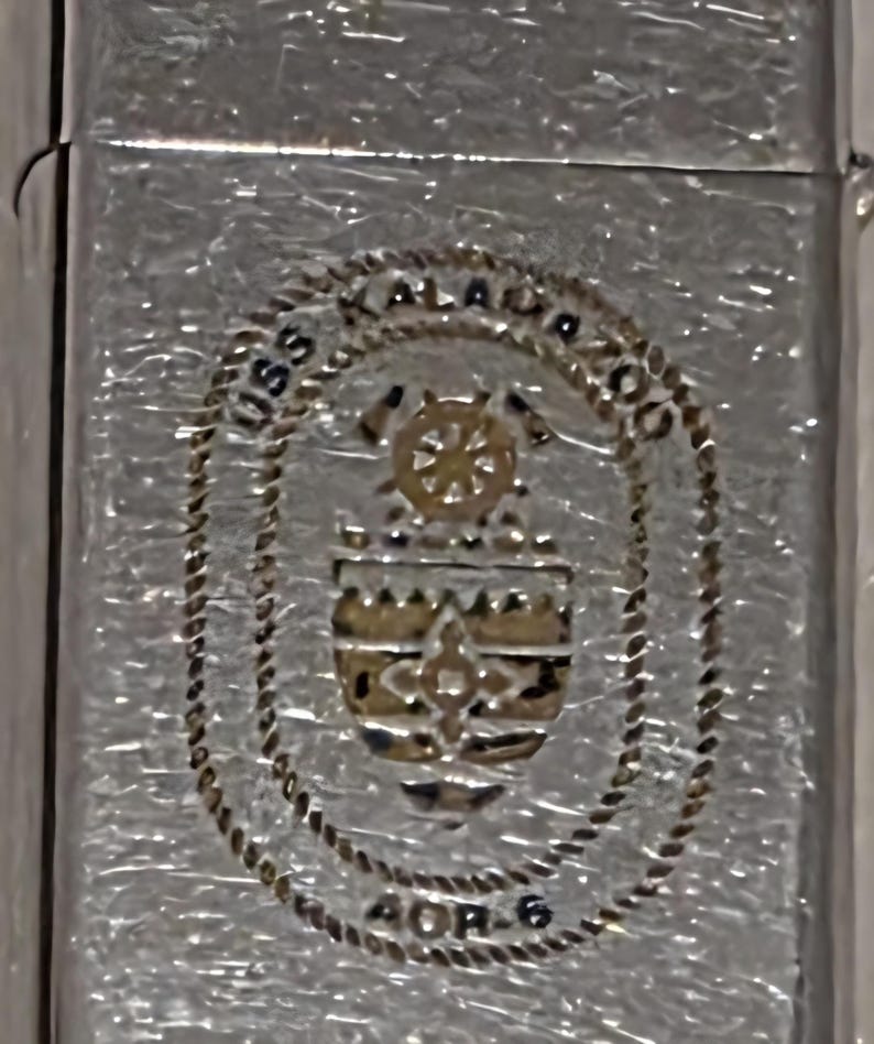 May include: Silver-toned metal lighter with a textured surface. The lighter features a detailed emblem with the text "USS KALAMAZOO" and "AOR-6" in a circular design. The emblem includes a ship's wheel and other nautical elements.
