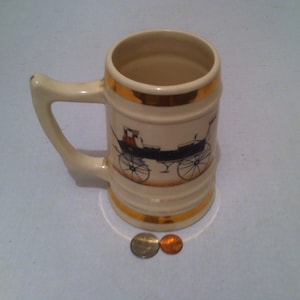 May include: A white ceramic beer stein with a gold rim and a black and white image of a horse-drawn carriage. The stein is sitting on a white surface.
