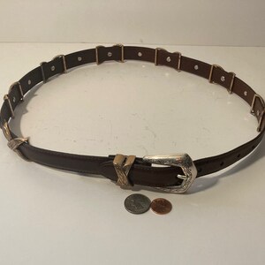 Puede incluir: Un cintur&oacute;n de cuero marr&oacute;n con una hebilla plateada y detalles dorados. La hebilla tiene un dise&ntilde;o intrincado y el cintur&oacute;n tiene m&uacute;ltiples bucles dorados.