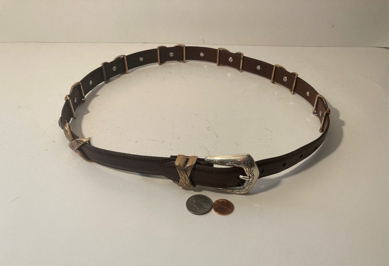 Puede incluir: Un cintur&oacute;n de cuero marr&oacute;n con una hebilla plateada y detalles dorados. La hebilla tiene un dise&ntilde;o intrincado y el cintur&oacute;n tiene m&uacute;ltiples bucles dorados.