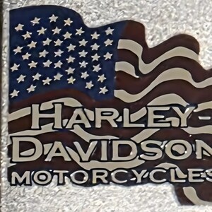 May include: A metal emblem featuring the American flag design with the text "HARLEY-DAVIDSON MOTORCYCLES". The flag has a blue field with white stars and red and white stripes. The text is in a bold, white font.