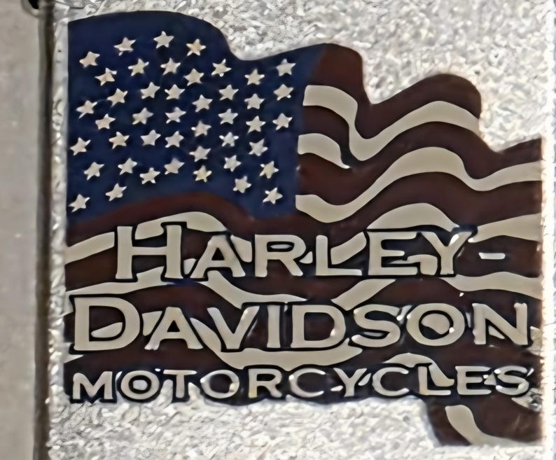 May include: A metal emblem featuring the American flag design with the text "HARLEY-DAVIDSON MOTORCYCLES". The flag has a blue field with white stars and red and white stripes. The text is in a bold, white font.