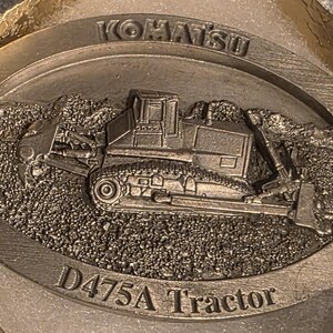 Op de afbeelding: Een zilverkleurig Komatsu D475A Tractor embleem. Het embleem toont een gedetailleerd reli&euml;f van een bulldozer op een getextureerde achtergrond, met het Komatsu-logo en de tekst "D475A Tractor".