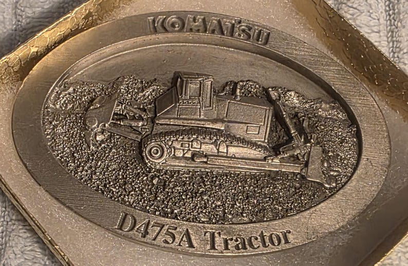Op de afbeelding: Een zilverkleurig Komatsu D475A Tractor embleem. Het embleem toont een gedetailleerd reli&euml;f van een bulldozer op een getextureerde achtergrond, met het Komatsu-logo en de tekst "D475A Tractor".