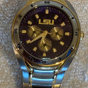 May include: Silver and gold-toned wristwatch with a purple face. The watch features the letters "LSU" in gold, three sub-dials, and a date display. The watch is on a silver and gold-toned metal band. Two coins are in the foreground.