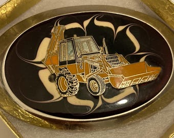 Puede incluir: Hebilla de cinturón ovalada con un diseño de vehículo de construcción en naranja y negro. El vehículo se encuentra sobre un fondo negro con remolinos crema, enmarcado por un borde plateado. La hebilla se presenta en una caja dorada.
