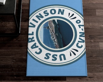Puede incluir: Una alfombra rectangular azul con un emblema circular con el texto "CARL VINSON" y "CVN-70 USS". El emblema contiene una imagen de un barco en el mar. La alfombra está sobre un suelo de madera oscura.