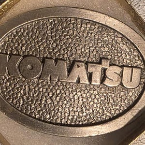 Op de afbeelding: Ovale metalen riemgesp met het woord "KOMATSU" in verhoogde letters. De achtergrond heeft een textuur, moza&iuml;ekachtig patroon. De gesp wordt gepresenteerd in een goudkleurige doos.