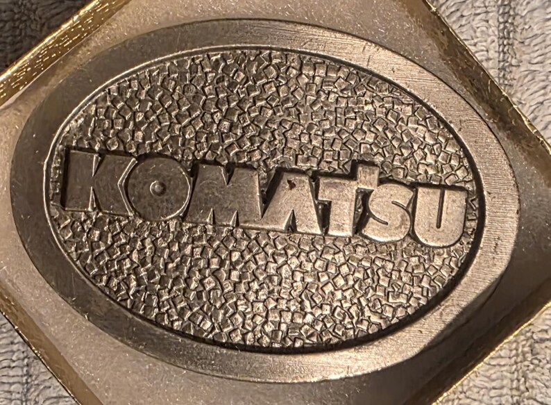 Op de afbeelding: Ovale metalen riemgesp met het woord "KOMATSU" in verhoogde letters. De achtergrond heeft een textuur, moza&iuml;ekachtig patroon. De gesp wordt gepresenteerd in een goudkleurige doos.