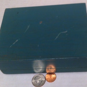 May include: A teal blue wooden box with a hinged lid. The box is slightly worn and has a few scratches on the surface. Two coins are sitting in front of the box.