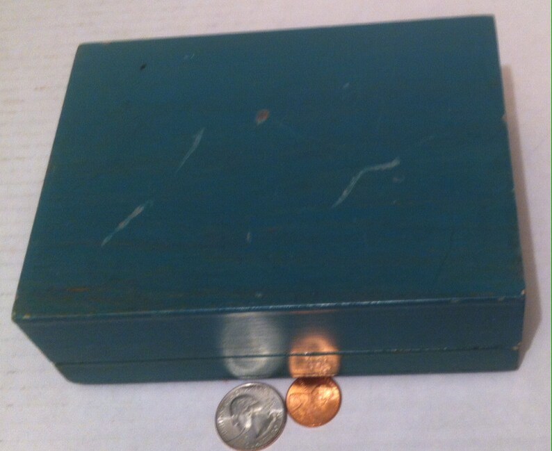 May include: A teal blue wooden box with a hinged lid. The box is slightly worn and has a few scratches on the surface. Two coins are sitting in front of the box.