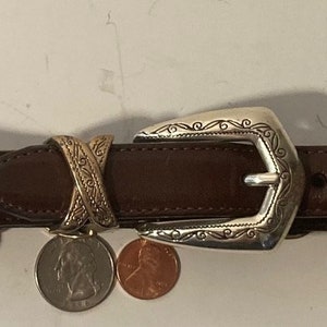 Puede incluir: Un cintur&oacute;n de cuero marr&oacute;n con una hebilla plateada y detalles dorados. La hebilla tiene un dise&ntilde;o intrincado y el cintur&oacute;n tiene un patr&oacute;n decorativo en X.