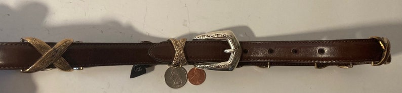 Puede incluir: Un cintur&oacute;n de cuero marr&oacute;n con una hebilla plateada y detalles dorados. La hebilla tiene un dise&ntilde;o intrincado y el cintur&oacute;n tiene un patr&oacute;n decorativo en X.