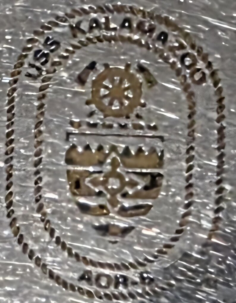 May include: A silver-toned emblem with the text "USS KALAMAZOO AOR-6" encircling a central design. The design includes a ship's wheel, a crest, and a cross-like symbol, all in a metallic gold color. The border has a rope-like texture.