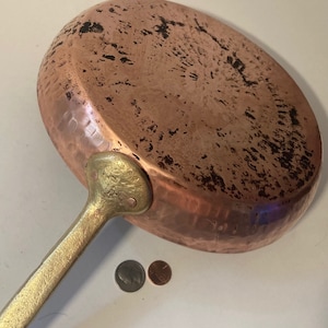 以下が含まれることがあります： ハンマー仕上げと真鍮製のハンドルが付いたビンテージの銅製のフライパン。フライパンは経年変化と使用による緑青があり、ハンドルはリベットで固定されています。フライパンは、スケールのために2つの日本のコインの隣に示されています。