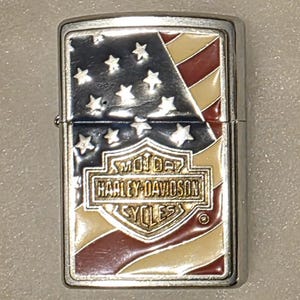 May include: A silver-colored Harley-Davidson lighter with an American flag design. The lighter features a red, white, and blue color scheme with the Harley-Davidson logo prominently displayed.
