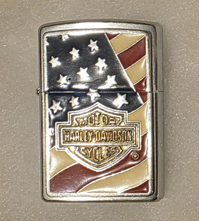 May include: A silver-colored Harley-Davidson lighter with an American flag design. The lighter features a red, white, and blue color scheme with the Harley-Davidson logo prominently displayed.