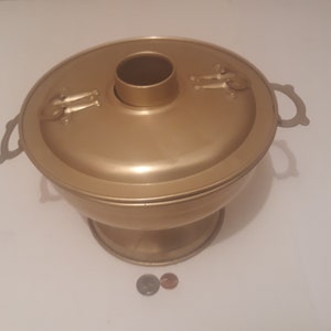 May include: A gold-colored metal hot pot with two handles and a lid. The pot has a round base and a raised center section with a hole in the top. The lid has a decorative design on the top.