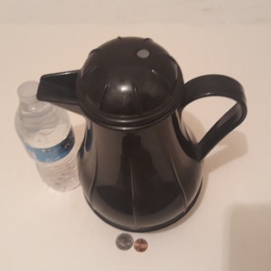 May include: A black thermal carafe with a lid and a handle. The carafe is sitting on a white surface with two coins next to it.