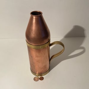 May include: A copper and brass bottle with a handle. The bottle has a rounded top and a cylindrical body. The bottle is decorated with a brass band around the top and bottom.