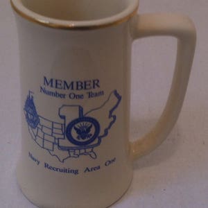 May include: White ceramic mug with a gold rim and blue and white graphic of a map of the United States with the text "MEMBER Number One Team Navy Recruiting Area One"