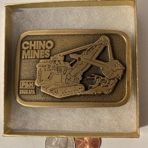 Peut inclure: Une boucle de ceinture dorée représentant une pelle minière P&H 2800 XPA détaillée avec les mots "Chino Mines" au-dessus.