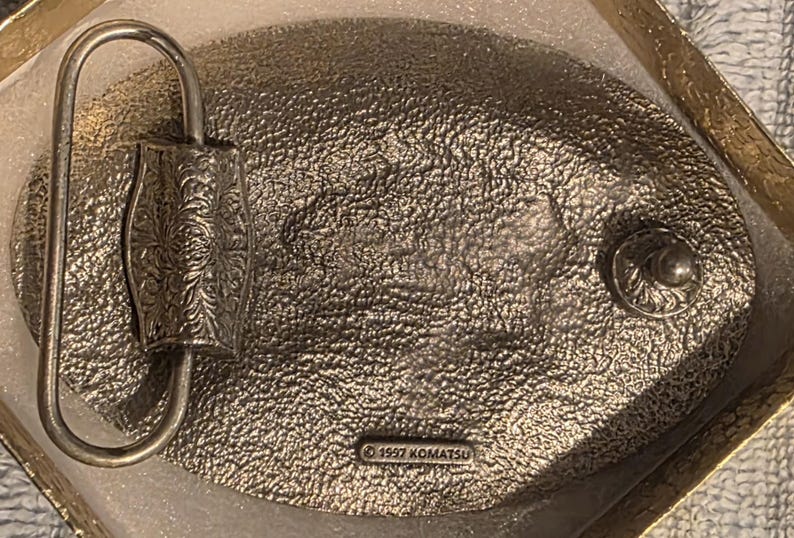 Op de afbeelding: Zilverkleurige metalen riemgesp met een ovale vorm en een getextureerd oppervlak. De gesp heeft een decoratief bloemmotief op de sluiting en de tekst "&copy; 1997 KOMATSU".