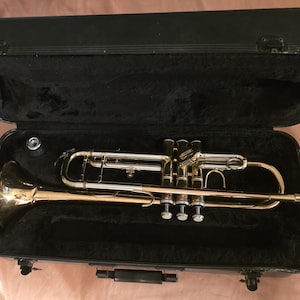 May include: A brass trumpet with silver accents rests inside an open black case with a plush, black interior. The trumpet's bell is a shiny gold color, and the valves and tubing are silver. The case is rectangular with a handle and latches.