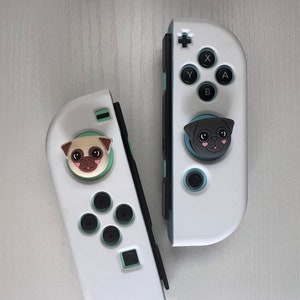 May include: Two white Nintendo Switch Joy-Con controllers with custom face buttons. The left controller has a pug face button and the right controller has a black pug face button.