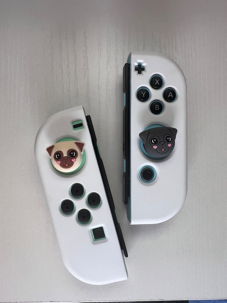 May include: Two white Nintendo Switch Joy-Con controllers with custom face buttons. The left controller has a pug face button and the right controller has a black pug face button.
