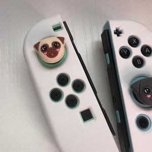 May include: Two white Nintendo Switch Joy-Con controllers with custom face buttons. The left Joy-Con has a green and white pug face button. The right Joy-Con has a black and white pug face button.