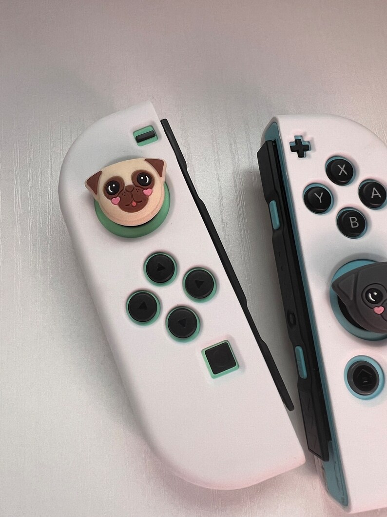 May include: Two white Nintendo Switch Joy-Con controllers with custom face buttons. The left Joy-Con has a green and white pug face button. The right Joy-Con has a black and white pug face button.