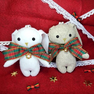 May include: Two plush bunny ornaments. One is white, the other is beige. Both have black eyes, a plaid bow, and a small gold bell. They are on a red background with white lace trim and gold star accents.