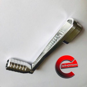 May include: A silver metal hand crank with a textured grip and a flat, rectangular handle. The handle has a small hole at the end. The crank is designed for use with a machine or device.